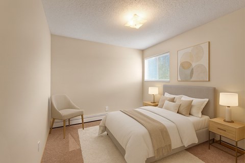 A cozy bedroom here at Aspen Terrace with beige carpet, soft neutral walls, and a gray upholstered bed dressed in crisp white bedding and a warm tan throw. A window with blinds brings in natural light, and matching nightstands with modern lamps create a calm, inviting feel.