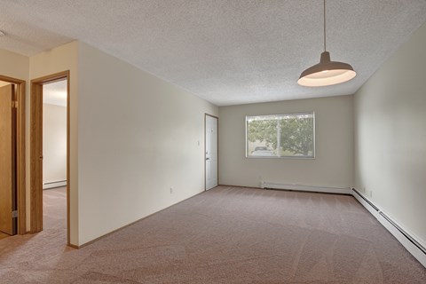 A spacious, empty living room here at Birch Terrace with soft beige carpet, neutral walls, and a modern hanging light fixture. A large window with blinds brings in natural light, and the open layout connects to a hallway leading to additional rooms.