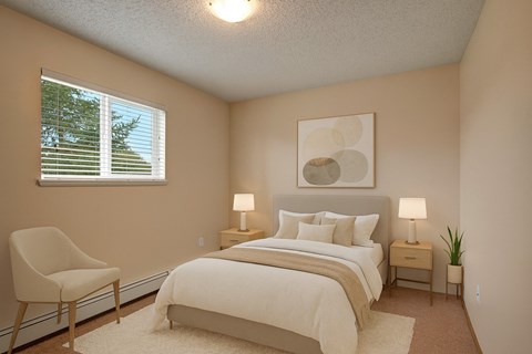 A cozy bedroom here at Birch Terrace with plush beige carpet, a soft neutral color palette, and a window with blinds for natural light. A comfortable bed with matching nightstands, warm lamps, and simple wall art creates a calm, inviting feel.