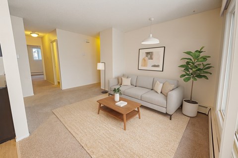 A bright, inviting living room here at Broadmoor with soft beige carpet, a light gray sofa with neutral pillows, and a warm wood coffee table on a textured area rug. A modern pendant light, simple wall art, and a tall green plant add a cozy, airy feel near large windows.