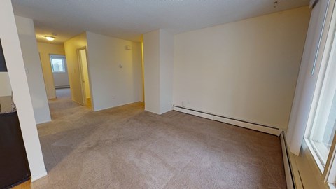 A spacious living area here at Broadmoor with soft carpet flooring, neutral walls, and a wide window that brings in natural light. A long baseboard heater runs along the wall, and the open layout connects smoothly to the hallway for an easy, comfortable flow.