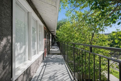 A sunny outdoor balcony here at Broadmoor with a wide balcony-style path, black metal railings, and large windows along the exterior wall. Mature green trees surround the view, creating a leafy, peaceful setting with plenty of natural shade and fresh air.
