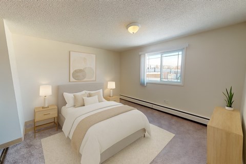 A bright bedroom here at Cambridge West with a soft upholstered bed, crisp white bedding, and matching nightstands with warm table lamps. A large window brings in natural light, while neutral carpeting, simple wall art, and a light wood dresser with a small plant create a calm, comfortable space.