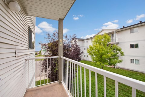 A private outdoor balcony here at Carrington Place with sturdy white railing and an open view of the community grounds, featuring lush green landscaping, mature trees, and a bright blue sky for a calm, refreshing outdoor escape.