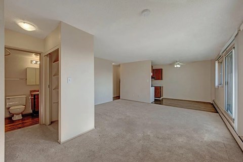 A spacious open living area here at Cedar Gardens with soft carpeting and an easy flow into the kitchen space, featuring large sliding glass doors with wide windows for natural light, a long baseboard heater, and nearby built-in shelving and bathroom access for added everyday convenience.