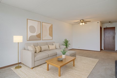 A bright living room here at Centre Pointe Plaza featuring a spacious light gray sectional with neutral throw pillows, a warm wood coffee table, and a soft area rug, complemented by simple wall art, a modern floor lamp, and fresh greenery for a cozy, welcoming feel.