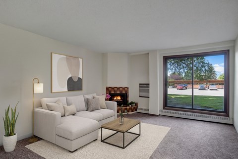 A cozy living room here at Edgemont Heights featuring a soft light gray sectional with neutral accent pillows, a warm wood coffee table on a textured area rug, and a glowing fireplace, complemented by simple wall art, greenery, and a large picture window with peaceful outdoor views.