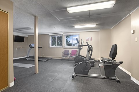 A bright fitness room here at Edgemont Heights featuring cardio equipment like a treadmill, elliptical, and stationary bike, plus open floor space for stretching or strength training, all in a clean, simple setting with windows for natural light.