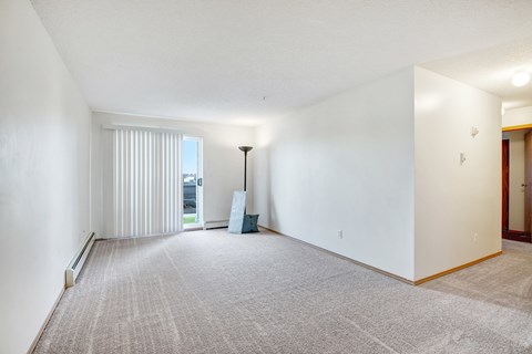 A bright living room here at Edgewood Estates featuring soft neutral carpet, large sliding patio doors with vertical blinds for natural light, and an open layout with plenty of space to relax, plus a simple floor lamp and minimal decor for a clean, welcoming feel.