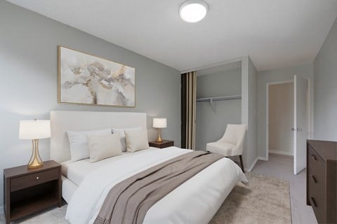 A cozy bedroom here at Gateway Gardens featuring a comfortable bed with crisp white bedding and neutral pillows, paired with matching nightstands and warm table lamps, plus simple wall art, an accent chair, and an open closet for easy storage.