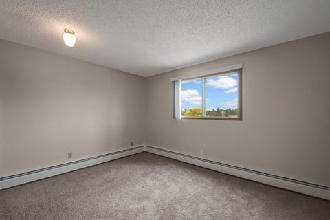 A bright, empty bedroom here at Gateway Square with soft beige walls, plush carpet flooring, and a large window with vertical blinds letting in natural light and peaceful outdoor views.