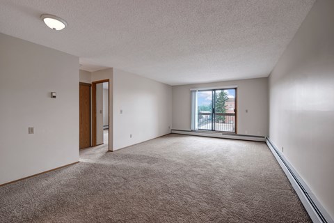 A bright living room here at Gateway Square featuring soft carpeting, crisp light walls, and a large sliding glass door that opens to a private balcony, filling the space with natural light and an airy feel.