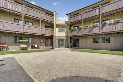 A welcoming courtyard exterior here at Kingsmere with multi-level apartment buildings, private balconies, landscaped grass, and a central walkway leading to a secure building entrance under a bright blue sky.