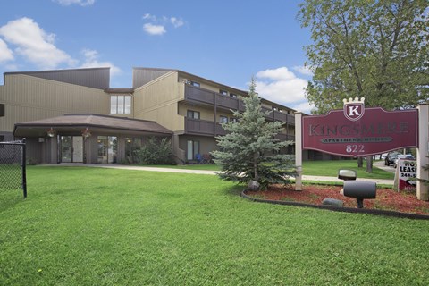 A welcoming exterior view here at Kingsmere with a landscaped green lawn, mature trees, a prominent community sign, and multi-level apartment buildings with balconies under a bright, sunny sky.
