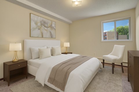 A softly staged bedroom here at Kingsmere with neutral walls, plush carpet, a comfortable bed with layered bedding, warm accent lighting, and a window bringing in natural light for a calm, inviting atmosphere.
