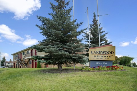 A welcoming Lakewood Heights community entrance sign with colorful landscaping, mature evergreen trees, and a well-maintained lawn that creates a friendly, residential vibe.