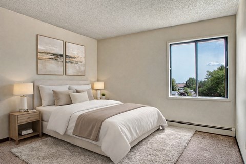 A bright, calming bedroom here at Madison Park featuring a neatly made bed with white bedding, layered neutral pillows, and a soft throw, paired with matching nightstands and warm table lamps, framed by minimalist wall art, plush carpet, and a large window offering natural light and neighborhood views.