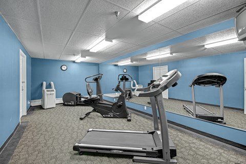 A well-equipped fitness center here at Madison Park with blue accent walls, mirrored panels, and carpeted flooring. The room features treadmills, stationary exercise bikes, and cardio equipment arranged for easy movement, while overhead lighting and wall mirrors create a bright, functional space for daily workouts.