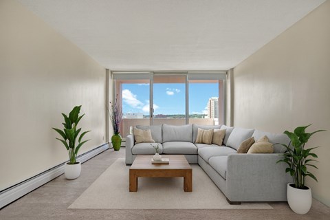 A bright, inviting living room here at Madison Park with soft neutral walls, beige carpet, and large sliding glass doors that fill the space with natural light and city views. A light gray sectional sofa with neutral pillows sits on an area rug, paired with a wooden coffee table and potted plants, creating a comfortable, modern setting.