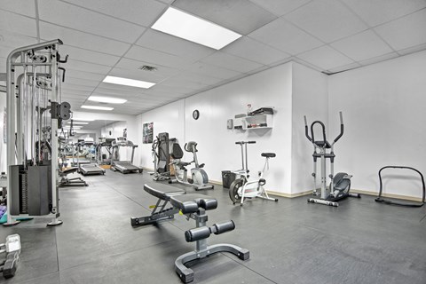 A spacious fitness center here at Madison Park with a variety of cardio and strength training equipment arranged across durable flooring. Treadmills, exercise bikes, weight machines, and free weights are visible, complemented by bright overhead lighting, mirrors, and clean white walls that create an open, motivating workout environment.