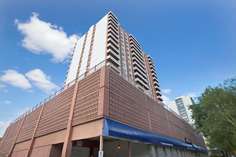A modern high-rise apartment building here at Madison Park showcasing a distinctive architectural design with stacked balconies, a textured exterior facade, and a prominent podium level below the residential towers, set against a bright blue sky with scattered clouds and surrounding greenery.