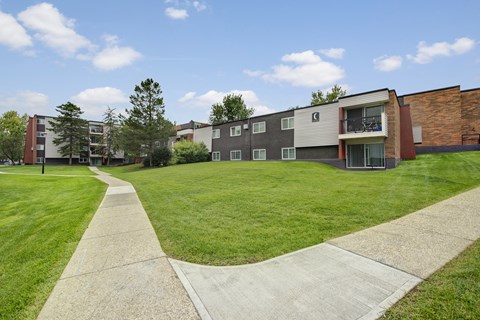 A spacious and well maintained outdoor courtyard here at Park Place South featuring expansive green lawns, curved concrete walking paths, mature trees, and multi level apartment buildings with balconies under a bright blue sky, creating a welcoming and walkable residential community setting.