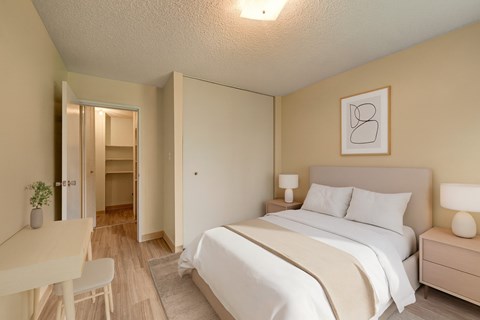 A cozy, modern bedroom here at Park Place South featuring a neatly made bed with soft neutral bedding, matching bedside tables with lamps, warm wood-style flooring, and a large closet, with light walls and simple decor creating a calm, comfortable space for rest and everyday living.