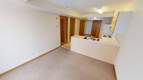 A bright open-concept living area here at Quail Ridge with soft carpet, neutral walls, and a wide breakfast bar with a stainless steel sink. A clean kitchen space features light cabinetry, a white refrigerator, and an electric range, creating an airy and functional layout.