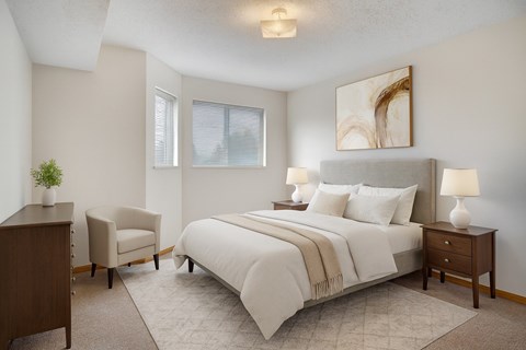 A bright bedroom here at Quail Ridge with soft carpet, neutral walls, and a cozy bed dressed in crisp white bedding and warm accents. A pair of nightstands with matching lamps and a cushioned chair create a calm, inviting space with gentle natural light from the windows.