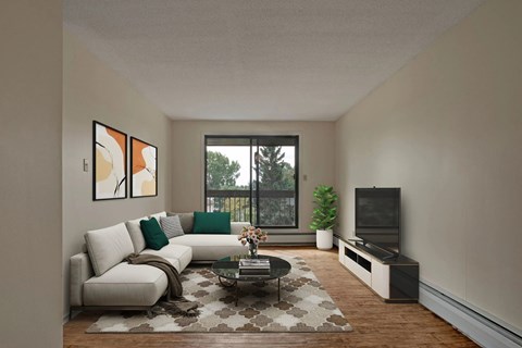 A bright living room here at Ranchlands featuring a light sectional sofa with accent pillows, abstract wall art, a patterned area rug, a round coffee table, and a media console with TV, complemented by wood-style flooring, neutral walls, and a sliding glass door leading to a private balcony with tree views.