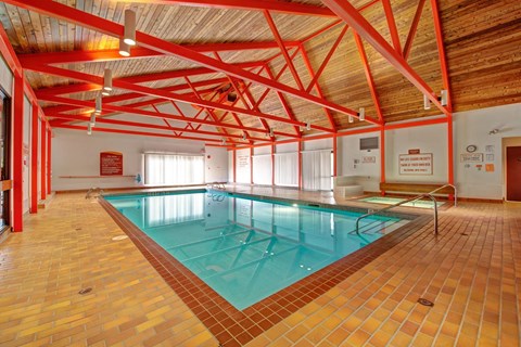 A spacious indoor swimming pool here at Ranchlands showcasing a rectangular lap pool with clear water, tiled pool deck, metal handrails, and safety signage, set beneath a vaulted wood ceiling with exposed red beams and ample overhead lighting that creates an open, recreational atmosphere.