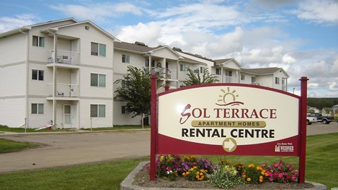 A welcoming exterior entrance sign here at Sol Terrace featuring the Sol Terrace Apartment Homes rental centre monument sign framed by colorful flower beds, with a three-story apartment building, balconies, paved drive, and neatly maintained green lawn visible in the background beneath a partly cloudy sky.