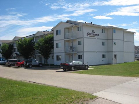 A landscaped exterior view here at Sol Terrace showcasing a three-story apartment building with light-colored siding, private balconies, surrounding green lawn areas, mature trees, and convenient on-site parking along a paved drive under a clear blue sky.