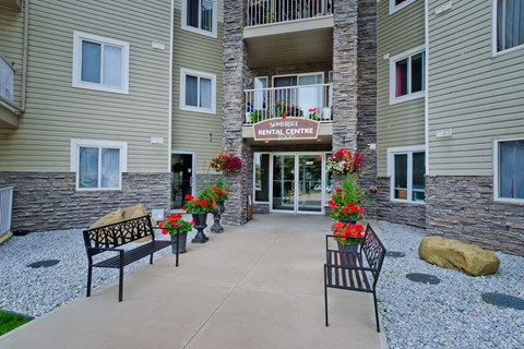 A welcoming exterior entrance here at Somerset Pointe featuring the rental centre doorway framed by stone accents, beige siding, and colorful flower planters, with benches along the concrete walkway and neatly landscaped gravel beds creating an inviting apartment community arrival.