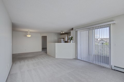 A bright open living room here at Somerset Pointe featuring soft neutral carpet, light gray walls, and a spacious layout that connects to a partial kitchen pass-through, highlighted by modern ceiling lighting and a large sliding glass door with vertical blinds that opens to a private balcony and brings in abundant natural light.