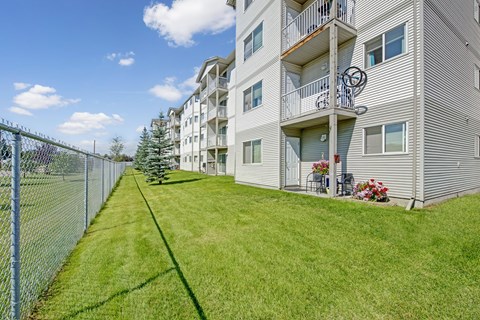 A landscaped exterior view here at Sonora showcasing a well-maintained green lawn alongside a multi-story apartment building with light-colored siding, private balconies on each level, patio seating with potted flowers, mature trees, and a fenced perimeter under a bright blue sky.