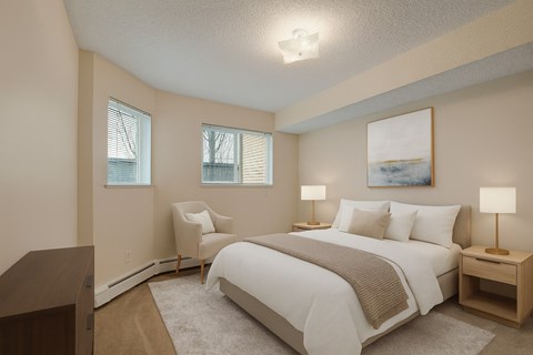 A cozy, light-filled bedroom here at Sonora featuring a queen-size bed with crisp white bedding, a soft neutral throw, matching bedside tables with warm lamps, plush carpet flooring, a comfortable accent chair, and windows with blinds that bring in natural light for a calm, inviting retreat.