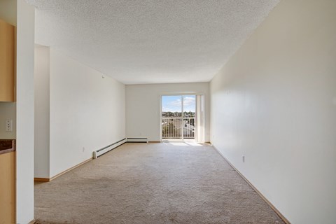 A bright and open living room here at Sonora featuring soft neutral carpeting, light cream walls, and a long rectangular layout that leads to a sliding glass door with vertical blinds opening to a private balcony, with ample natural light, a baseboard heater, and clean finishes creating a flexible and inviting space.