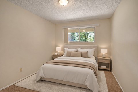 A cozy bedroom here at Stirling Place featuring a queen-sized bed with neutral bedding and layered pillows, matching nightstands with soft table lamps, plush carpet flooring, light beige walls, and a window above the headboard that brings in natural light and a peaceful outdoor view.