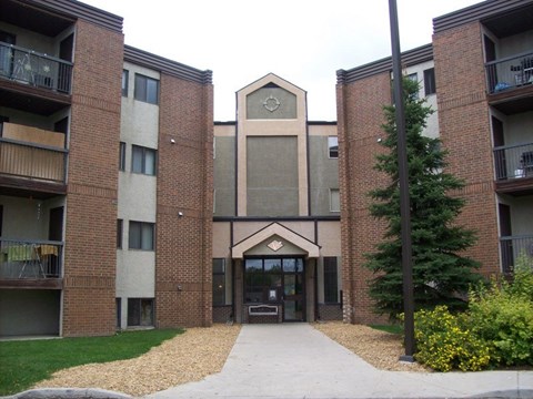 A welcoming apartment building exterior here at Ventana showcasing a multi-story brick and neutral-tone façade, a central covered entrance with glass doors, private balcony railings on each level, landscaped walkways, and mature greenery that create a well-maintained and inviting community setting.