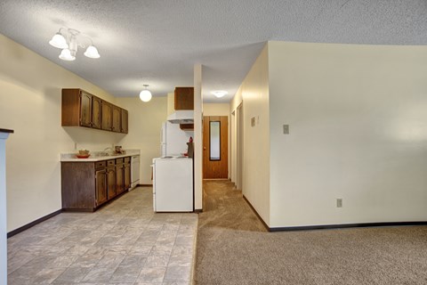 A bright open living space here at Ventana showcasing a combined living and kitchen layout with neutral walls, soft carpet and tile flooring, wood-style cabinetry, white appliances, overhead lighting, and a spacious hallway that creates a functional, comfortable flow throughout the apartment.