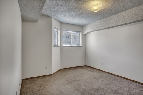 A bright bedroom here at Villagio showcasing soft neutral walls, plush carpet flooring, a unique ceiling beam detail, and two compact windows with blinds that bring in natural light, creating a comfortable and versatile space suitable for a variety of furniture layouts and everyday living.