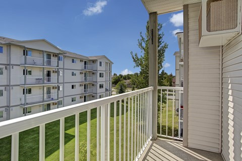 A bright private balcony here at Villagio overlooking landscaped green lawns and neighboring apartment buildings, featuring white railing, covered outdoor space, and open sky views that provide a relaxing spot for fresh air, outdoor seating, and enjoying the surrounding community scenery.