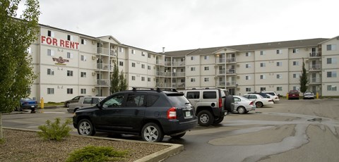 A wide exterior view here at Wellington Manor featuring a multi-story apartment community with private balconies and a large resident parking lot. A well-kept driveway and landscaped greenery create a welcoming first impression. A clear “For Rent” sign on the building makes it easy to spot the community from the street.