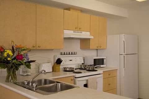 A bright kitchen here at Wellington Manor with light wood cabinets, clean white countertops, and a double stainless steel sink. A white stove with a vent hood, a microwave, and a full-size refrigerator provide a practical cooking setup. A wide breakfast bar counter adds extra prep space and a convenient spot for casual dining.