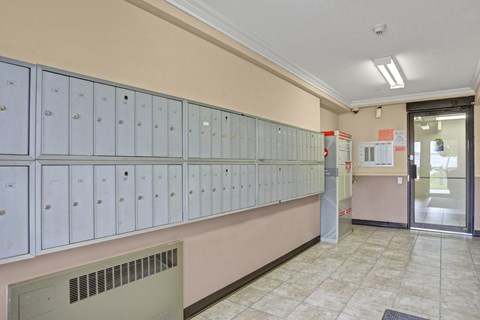 A clean community mailroom here at Wellington Manor with a long wall of secure resident mailboxes and bright overhead lighting. A tiled floor and neutral wall tones create a neat and well-kept space. A convenient entry door and parcel area provide easy access for everyday mail pickup.