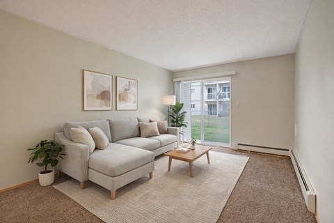 A bright living room here at Westhaven Estates with soft carpet flooring, a light gray sectional sofa, and a neutral area rug for a cozy feel. A sliding glass door lets in natural light and opens to a grassy outdoor view. A simple wooden coffee table, minimal wall art, and greenery accents add a clean and relaxing vibe.