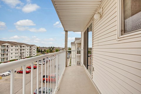 A private balcony here at Westhaven Estates with a covered concrete patio and white railing overlooking the community grounds. A sliding glass door provides easy indoor-outdoor access and plenty of natural light. A scenic view of nearby buildings and the parking area creates an open, airy feel.
