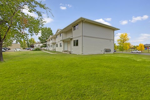 A peaceful outdoor view here at Westhaven Estates featuring a row of apartment buildings with light siding and private entry doors. A wide stretch of green lawn and mature trees creates a spacious setting, with a sidewalk connecting the homes to nearby parking and community areas. Bright blue skies and fall-colored trees add a cheerful touch to the landscape.