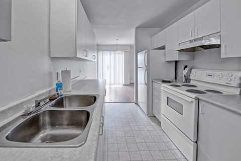 A bright galley-style kitchen here at Willowview Terrace with white cabinets, light countertops, and a double stainless steel sink. White appliances including a stove, range hood, and refrigerator line the space, with durable tile flooring that leads into the dining area with a sliding glass door for natural light.